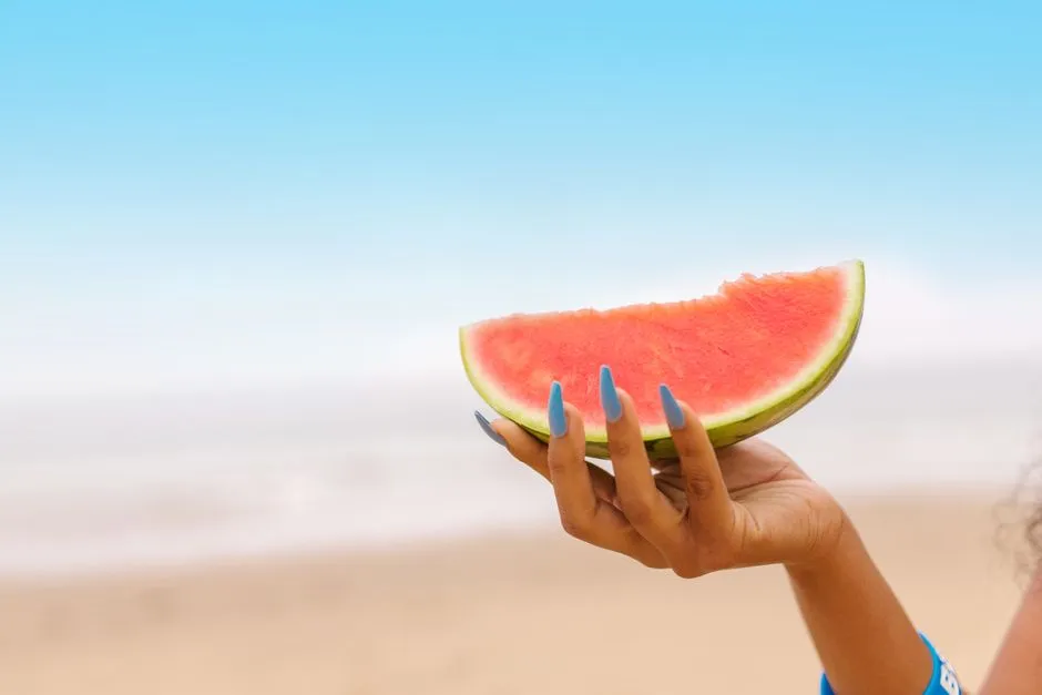 Hånd med lange løsnegle holder en melon på stranden, klar til at bade med løsnegle